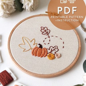 Può includere: Un modello PDF stampabile e istruzioni per un telaio da ricamo con un design a tema autunnale. Il design presenta una zucca, foglie e vortici in tonalità di arancione, marrone e giallo.