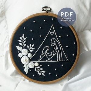Puede incluir: Arte de bordado en aro con un contorno blanco de la escena de la natividad con una estrella y un corazón sobre un fondo negro. El aro está rodeado de cuentas de perlas blancas y un diseño floral blanco.