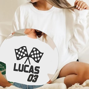 May include: White long-sleeved sweatshirt with a black and white chequered flag design, the name "LUCAS" and the number "03" embroidered in black. A casual style.