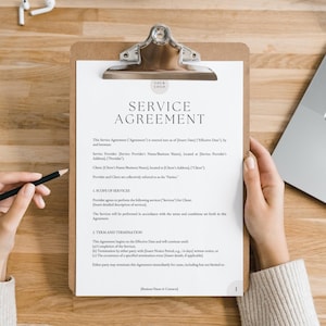 Peut inclure: Un presse-papiers blanc avec un document de contrat de service. Le document comprend le titre "Service Agreement" et des sections pour la port&eacute;e des services, la dur&eacute;e et la r&eacute;siliation, et le nom de l'entreprise en contact. Le document est sur une table en bois avec un ordinateur portable et un crayon.
