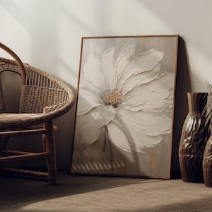 Puede incluir: Una impresión en lienzo enmarcada de una flor blanca sobre un fondo beige. La impresión se apoya contra una pared junto a una silla de mimbre y dos jarrones decorativos.