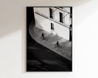 Op de afbeelding: Zwart-witfoto van een straathoek in de stad met twee mensen die fietsen. De straat is leeg en het gebouw op de achtergrond heeft een witte gevel met ramen.