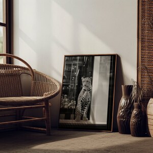 May include: A black and white photograph of a leopard in a window. The leopard is looking directly at the camera. The photograph is framed in a dark wood frame and is hanging on a white wall.