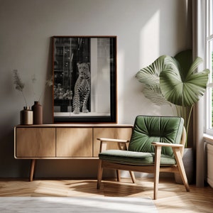 May include: A black and white photograph of a leopard in a window display, hanging above a wooden cabinet with three drawers and a green velvet armchair.