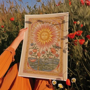 May include: A vintage-style print of a sun with a face surrounded by stylized flowers and leaves. The print is framed in a gold-colored frame and has a cream-colored background.