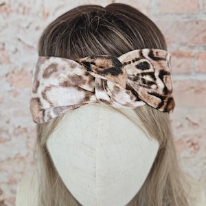 Könnte beinhalten: Ein braunes und weißes Stirnband mit Leopardenmuster und einem Knotendesign. Das Stirnband ist aus weichem, dehnbarem Stoff.