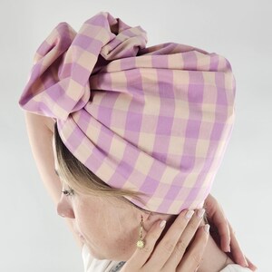 Könnte beinhalten: Ein Turban aus lila-beigem Karostoff. Der Stoff ist um den Kopf gewickelt und oben geknotet. Der Turban ist aus weichem Material, wahrscheinlich Baumwolle. Ein modisches Kopfbedeckungsaccessoire.