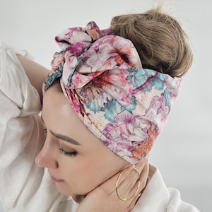 Könnte beinhalten: Ein Turban mit floralem Muster in Rosa, Lila und Türkis. Der Kopfschmuck hat ein großes, geknotetes Design und wird auf dem Kopf getragen. Der Stoff hat ein weiches, fließendes Aussehen. Ein goldener Creolen-Ohrring ist sichtbar.