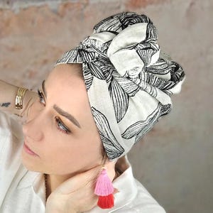 Könnte beinhalten: Ein weißer Turban mit schwarzem Blumenmuster. Der Turban ist oben auf dem Kopf zu einem Knoten gebunden. Die Person, die den Turban trägt, hat rosa Quastenohrringe.
