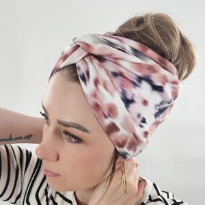 Könnte beinhalten: Ein gemustertes Stirnband in Weiß-, Mauve- und Lilatönen. Der Stoff ist oben verdreht und schafft so ein stilvolles Accessoire. Das Stirnband wird auf dem Kopf getragen, mit hochgestecktem Haar.