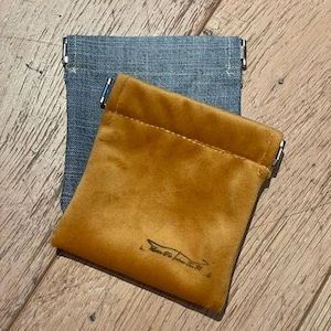 Op de afbeelding: Twee kleine zakjes. Eén is van blauwe denim, de andere van mosterdgeel fluweel. Het gele zakje heeft een klein zwart logo. Beide zakjes hebben zilveren metalen accenten aan de bovenkant.