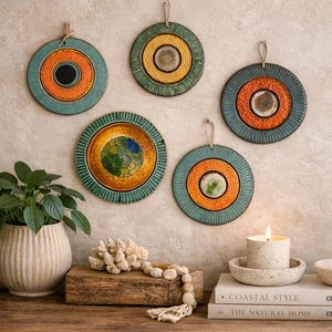 Puede incluir: Cinco adornos de pared de cerámica con diseños circulares en tonos naranja, turquesa y verde. Cada pieza tiene una superficie texturizada y un lazo de yute para colgar. La composición incluye una vela, libros y una planta.