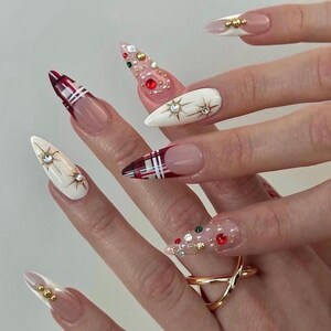 May include: A set of long, pointed fingernails with various designs. Some nails feature a clear base with red, green, and gold gems. Others have a white base with star-shaped embellishments or a red and white plaid pattern. A gold ring is also visible.