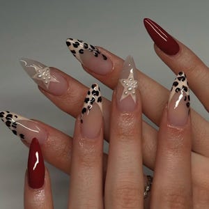 May include: Close-up of a hand with long, pointed nails. The nails are painted with a clear base and a leopard print design. Some nails have a white star with pearl accents.