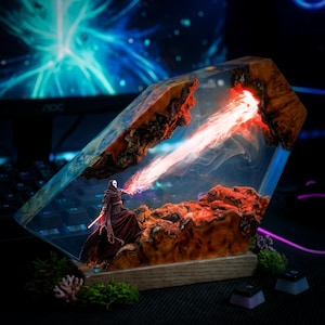 Op de afbeelding: Een harskunstwerk met een figuur met een rood lichtzwaard, dat een rode straal uitzendt. Het kunstwerk is ingekapseld in een helder harsblok met een houten basis, met een gedetailleerd landschap met rode en bruine elementen. De achtergrond heeft een blauw en zwart thema.