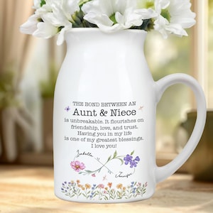 May include: White ceramic pitcher with a handle, filled with white flowers. The pitcher has text that reads "Aunt & Niece" and a message about their bond. Floral and butterfly accents decorate the pitcher.