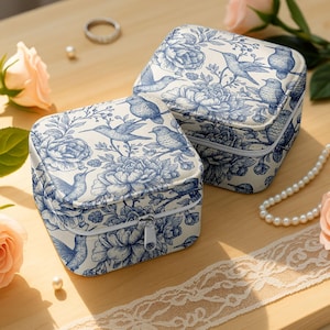 May include: Two square jewellery boxes with a blue and white floral and bird pattern. Each box has a white zip closure. The boxes are on a wooden surface with a pearl necklace and a pink rose.