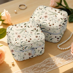 以下が含まれることがあります： クリーム色のジュエリーボックス2つ。花柄模様で、ピンク、茶色、緑の花と葉の繊細なデザイン。ボックスは正方形で、ジッパー開閉式。木製の表面に置かれています。