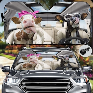 May include: A car sunshade featuring two cows, one wearing a pink headband and the other wearing aviator sunglasses. The sunshade is black with a gray interior.