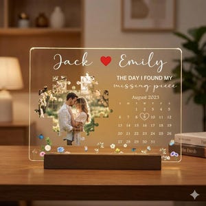 May include: A personalized acrylic photo display with a wooden base. The display features a photo puzzle, names "Jack" and "Emily", a heart, and the text "THE DAY I FOUND MY missing piece". It includes a calendar for August 2023.