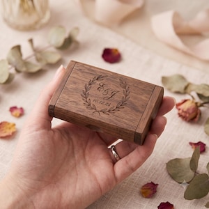 Puede incluir: Una caja rectangular de madera marrón oscuro con tapa abatible. La parte superior de la tapa presenta un diseño grabado con láser que consiste en una corona de laurel que rodea las letras E&J y la fecha 10.12.24 en fuente cursiva y serif.