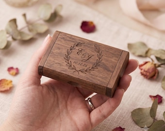 May include: A rectangular dark brown wooden box with a hinged lid. The top of the lid features a laser-engraved design consisting of a circular laurel wreath surrounding the letters E&J and the date 10.12.24 in a script and serif font.
