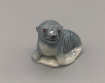 May include: A small ceramic figurine of a gray and white seal pup. The seal is lying on its belly with its head turned to the side. It has brown eyes and a small black nose.