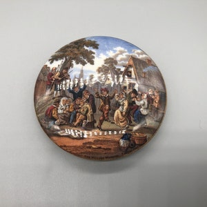 May include: A round ceramic lid with a colorful illustration of a village scene. The scene depicts people dancing and playing music in front of a building. The text "The Village Wedding" is printed on the lid.
