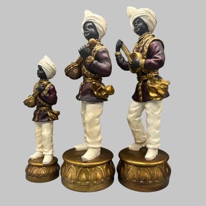 May include: Three decorative figurines of musicians. Each figure has a black face, white turban, and is dressed in a maroon and gold outfit. The figures stand on ornate gold bases. One figure plays a stringed instrument.