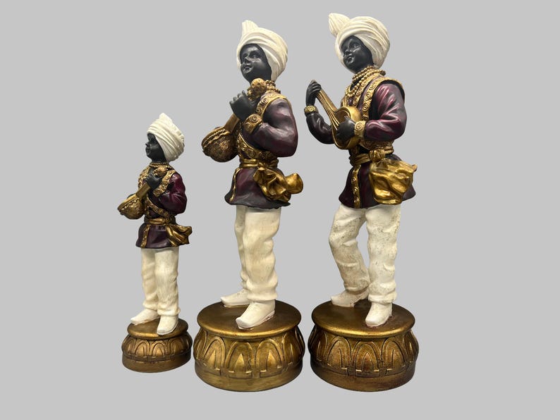 May include: Three decorative figurines of musicians. Each figure has a black face, white turban, and is dressed in a maroon and gold outfit. The figures stand on ornate gold bases. One figure plays a stringed instrument.