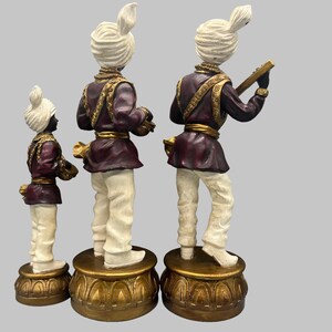 May include: Three decorative figurines of musicians. Each figure wears a white turban, a burgundy jacket with gold trim, and white pants. One figure holds a stringed instrument. The figurines stand on ornate gold bases.