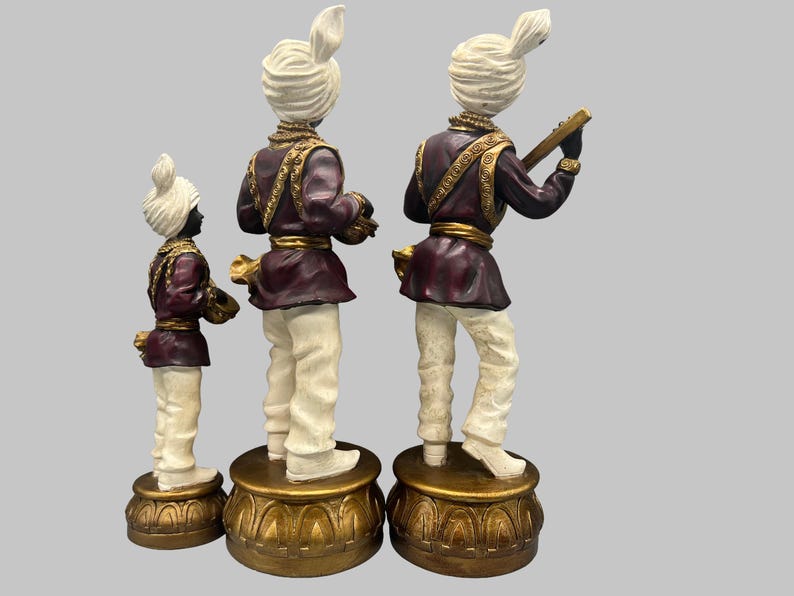 May include: Three decorative figurines of musicians. Each figure wears a white turban, a burgundy jacket with gold trim, and white pants. One figure holds a stringed instrument. The figurines stand on ornate gold bases.
