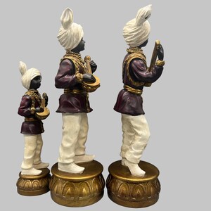 May include: Three decorative statues of musicians. Each figure wears a white turban, a maroon jacket with gold trim, and white pants. The statues stand on gold-colored pedestals and hold musical instruments.