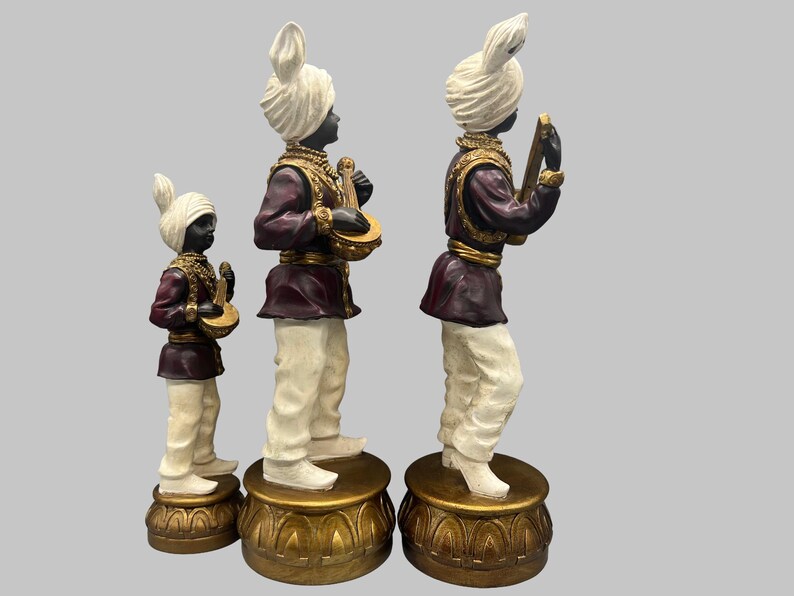 May include: Three decorative statues of musicians. Each figure wears a white turban, a maroon jacket with gold trim, and white pants. The statues stand on gold-colored pedestals and hold musical instruments.