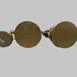 May include: Three antique brass-colored figurines with circular bases. The bases are different sizes, with the largest in the center. The figurines have white and brown details. The background is a solid light gray.