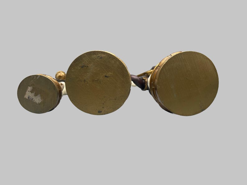May include: Three antique brass-colored figurines with circular bases. The bases are different sizes, with the largest in the center. The figurines have white and brown details. The background is a solid light gray.