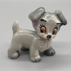 May include: A small ceramic figurine of a grey and white Dalmatian dog with black eyes and pink ears. The dog is standing on all four paws and has a playful expression on its face.