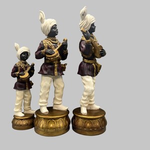 May include: Three decorative figurines of musicians. Each figure is dressed in a turban, maroon jacket, and white pants. They stand on gold-colored pedestals and hold musical instruments. The figures are black with white turbans.