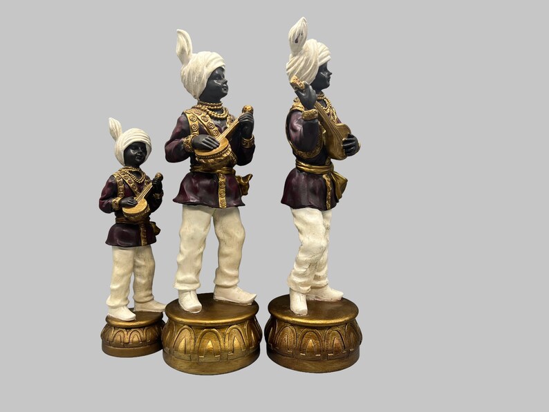 May include: Three decorative figurines of musicians. Each figure is dressed in a turban, maroon jacket, and white pants. They stand on gold-colored pedestals and hold musical instruments. The figures are black with white turbans.