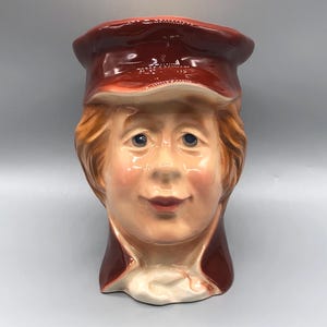 May include: A ceramic figurine of a person with a red hat and a brown jacket. The person has a friendly expression and is smiling.
