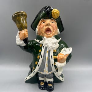 May include: A ceramic figurine of a town crier, dressed in a green coat with white trim and a black hat. The figurine holds a bell in one hand and a scroll in the other. The crier's face is animated with an open mouth.