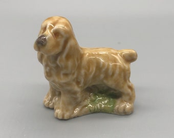 May include: A small ceramic figurine of a brown and white Cocker Spaniel dog. The figurine is standing on a green base with the word "WADE" printed on it.