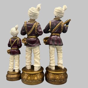 May include: Three decorative figurines of musicians in traditional attire. The figures are in a row, each standing on a gold-colored base. They wear white turbans, burgundy jackets with gold trim, and white pants. One figure holds a musical instrument.