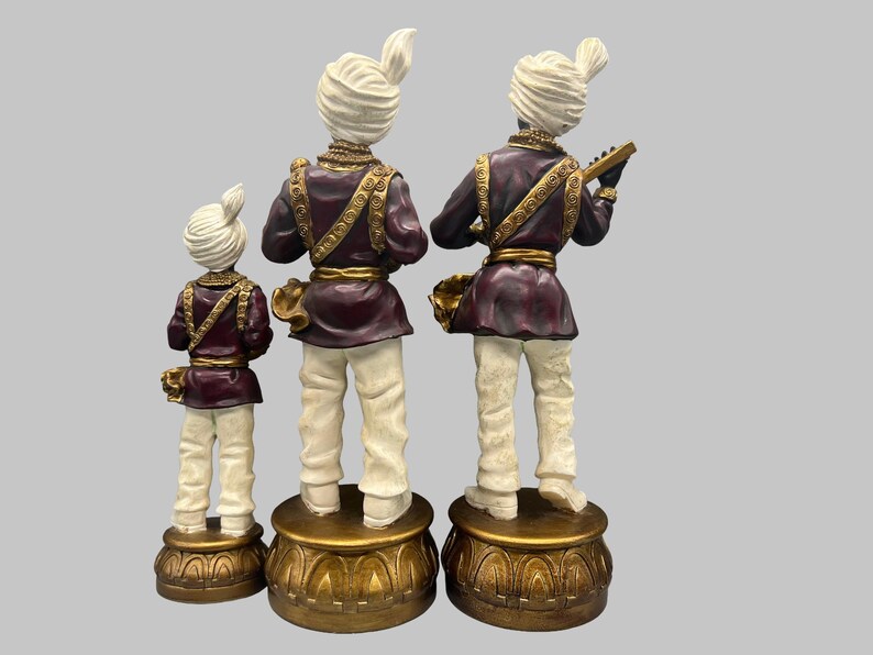 May include: Three decorative figurines of musicians in traditional attire. The figures are in a row, each standing on a gold-colored base. They wear white turbans, burgundy jackets with gold trim, and white pants. One figure holds a musical instrument.