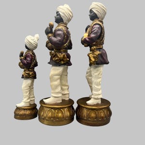 May include: Three decorative figurines of men in traditional attire. The figures have black skin, white turbans, and are dressed in purple and gold jackets, white pants, and stand on ornate gold bases. The figures are of varying heights.