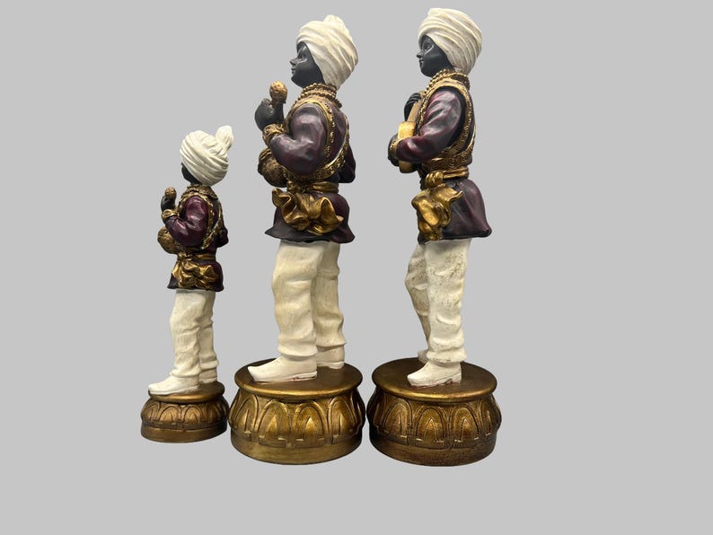 May include: Three decorative figurines of men in traditional attire. The figures have black skin, white turbans, and are dressed in purple and gold jackets, white pants, and stand on ornate gold bases. The figures are of varying heights.
