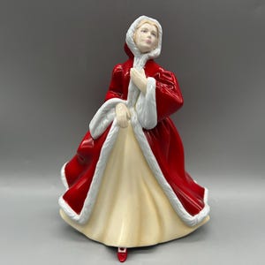 May include: A ceramic figurine of a woman in a red cloak with white trim and a cream-colored dress. The figurine has a hood and is posed with hands clasped. The base is cream-colored with a red foot.