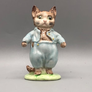 May include: A ceramic figurine of a cat wearing a light blue outfit. The cat has brown and tan fur, green eyes, and a serious expression. It stands on a small, oval base of green and yellow. The figurine is a decorative item.