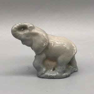 May include: A small, ceramic elephant figurine in shades of grey. The elephant is posed with its trunk raised and is standing on a base. The word "WADE" is visible on the base. The figurine has a glossy finish.