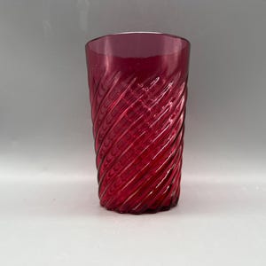 May include: A tall, ruby-red glass with a spiral design. The glass has a textured surface and a cylindrical shape. The colour is a deep, rich red, and the glass appears to be made of a thick material. The glass is empty and sits on a white surface.